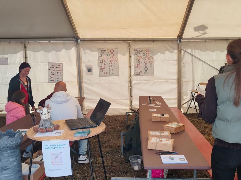 Sous une grande tente blanche, des animatrices discutent avec des enfants et des adultes. Sur les tables du stand, un ordinateur, une peluche de chamois, des boîtes cadenassés. Au fond, les affiches CLEFs de Phonocolor.